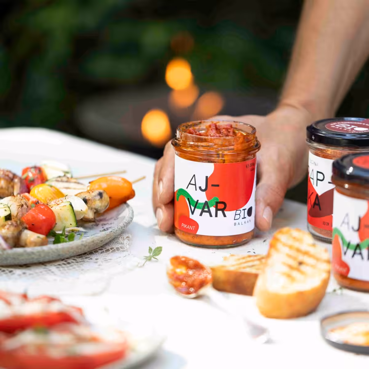 Ein Glas Bio Ajvar auf einem Tisch gedeckt mit Grillspießen, mit Feuer von einem Grill im Hintergrund